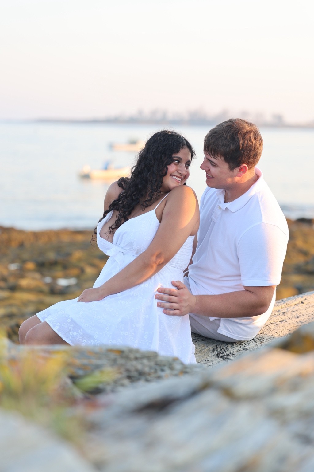 A smiling couple seated on driftwood beside the ocean